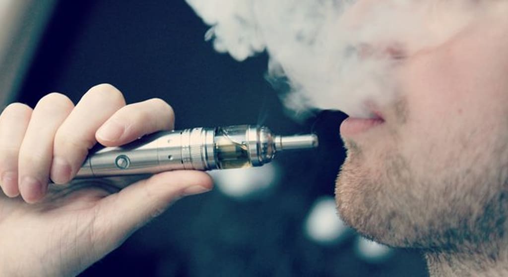 A man exhaling a large cloud of vapor from a vape pen held in his hand.
