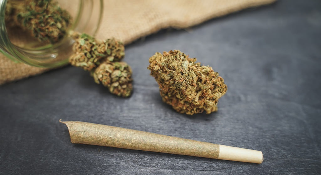A single cannabis bud and a pre-rolled joint on a dark grey surface, with a glass jar of buds and burlap in the background.