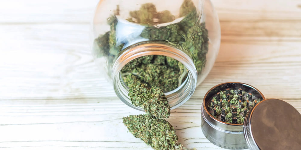 Green THCA cannabis buds spilling from an overturned glass jar onto a light wooden surface, next to an open metal grinder with ground cannabis.