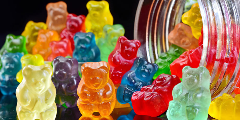Brightly colored gummy bears spilling out of a glass jar onto a reflective surface.