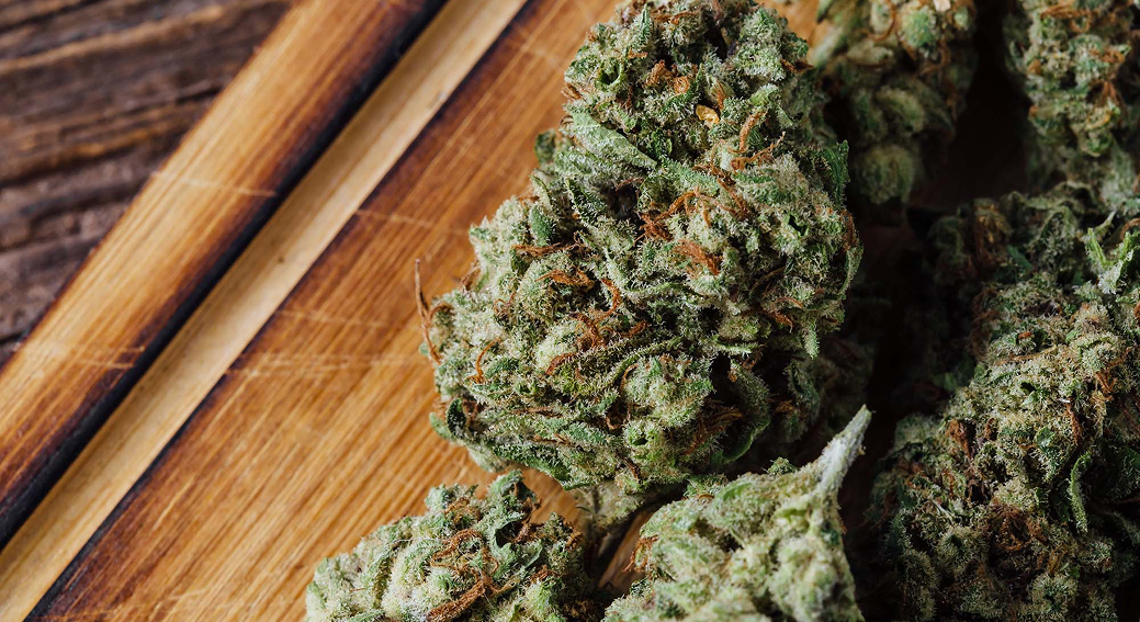 A macro shot of several dried and cured cannabis buds on a wooden cutting board, covered in a frosty layer of trichomes.