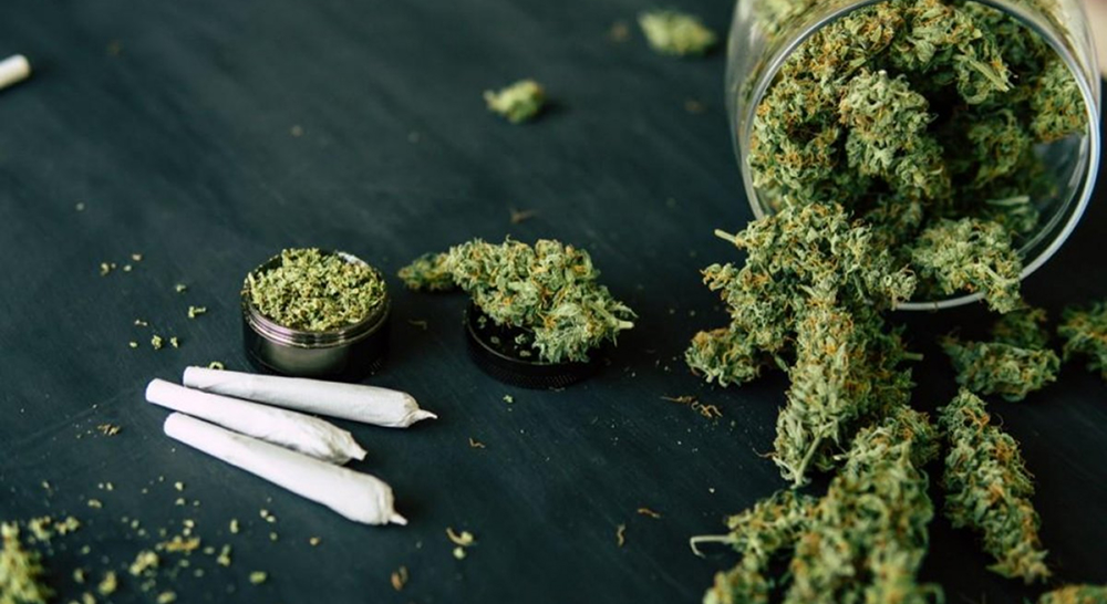 A flat-lay photo on a dark surface, showing a glass jar tipped over with cannabis buds spilling out. Next to it are three pre-rolled joints and a small metal grinder with ground-up cannabis inside.